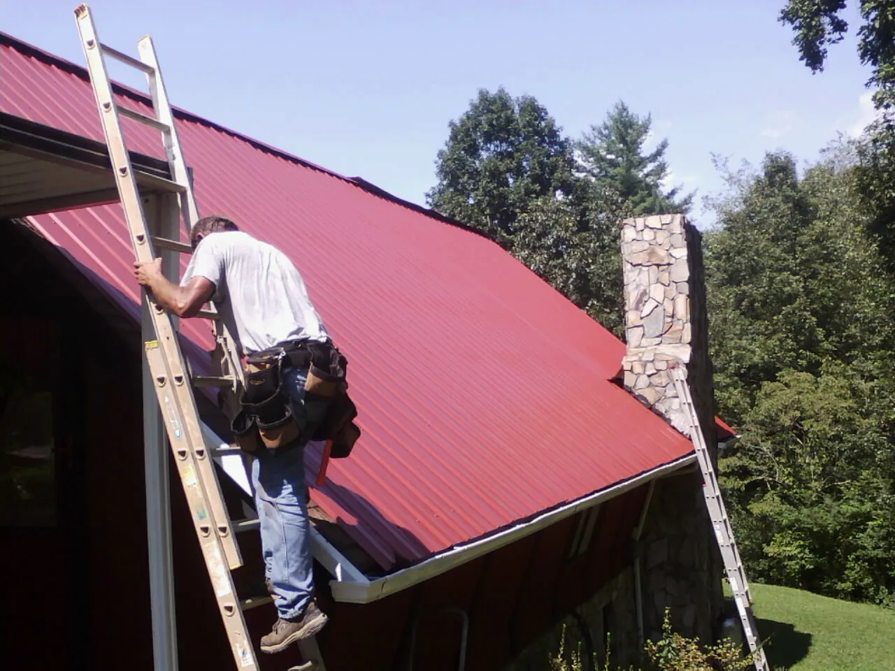 Roofer on a ladder beside a red metal roof and stone chimney — Soffit & Fascia Repair by Roofing Brothers in Hopewell
